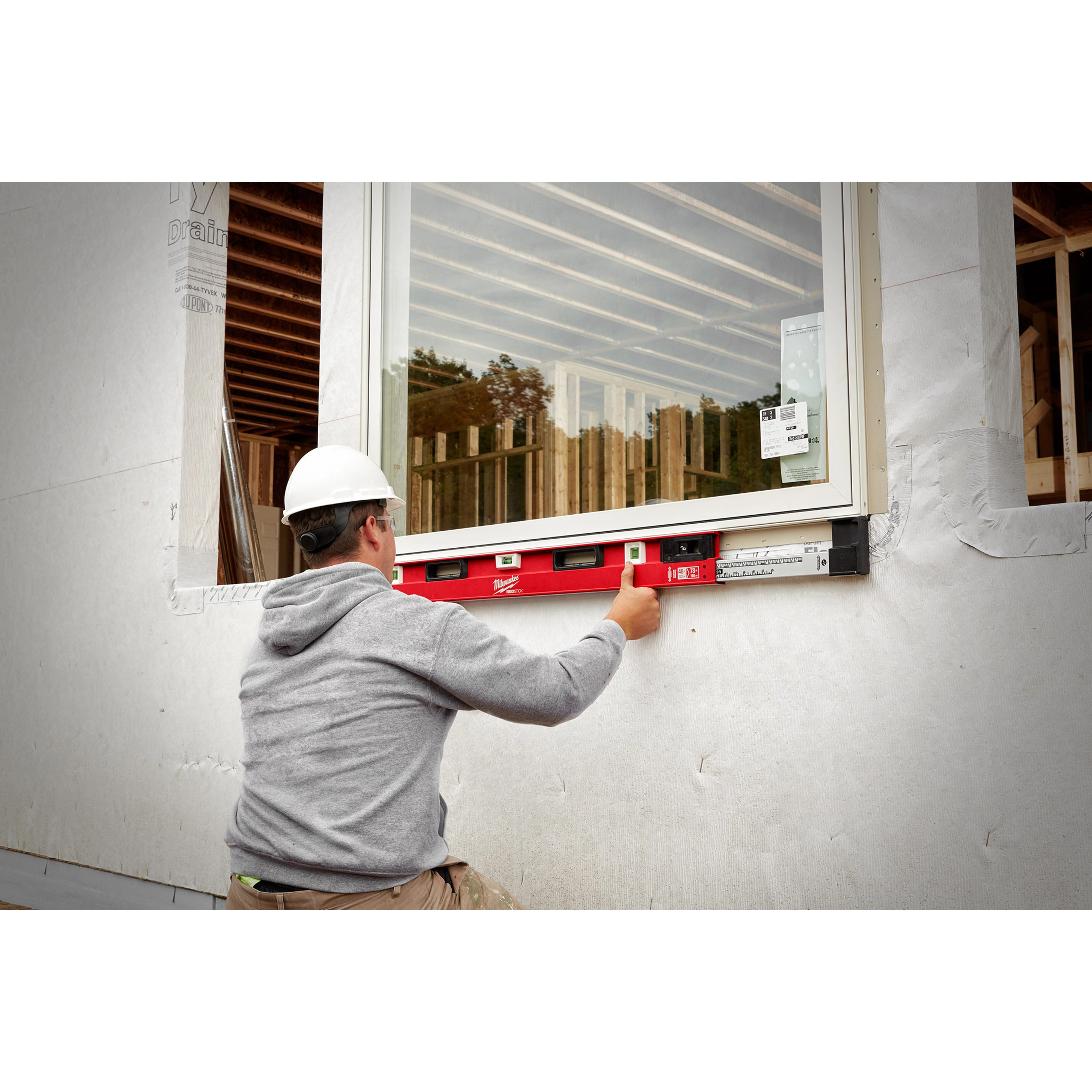 A construction worker in a gray hoodie uses a red 48"-78" Expandable Install Level to align a window during installation.