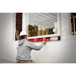 A construction worker in a gray hoodie uses a red 48"-78" Expandable Install Level to align a window during installation.