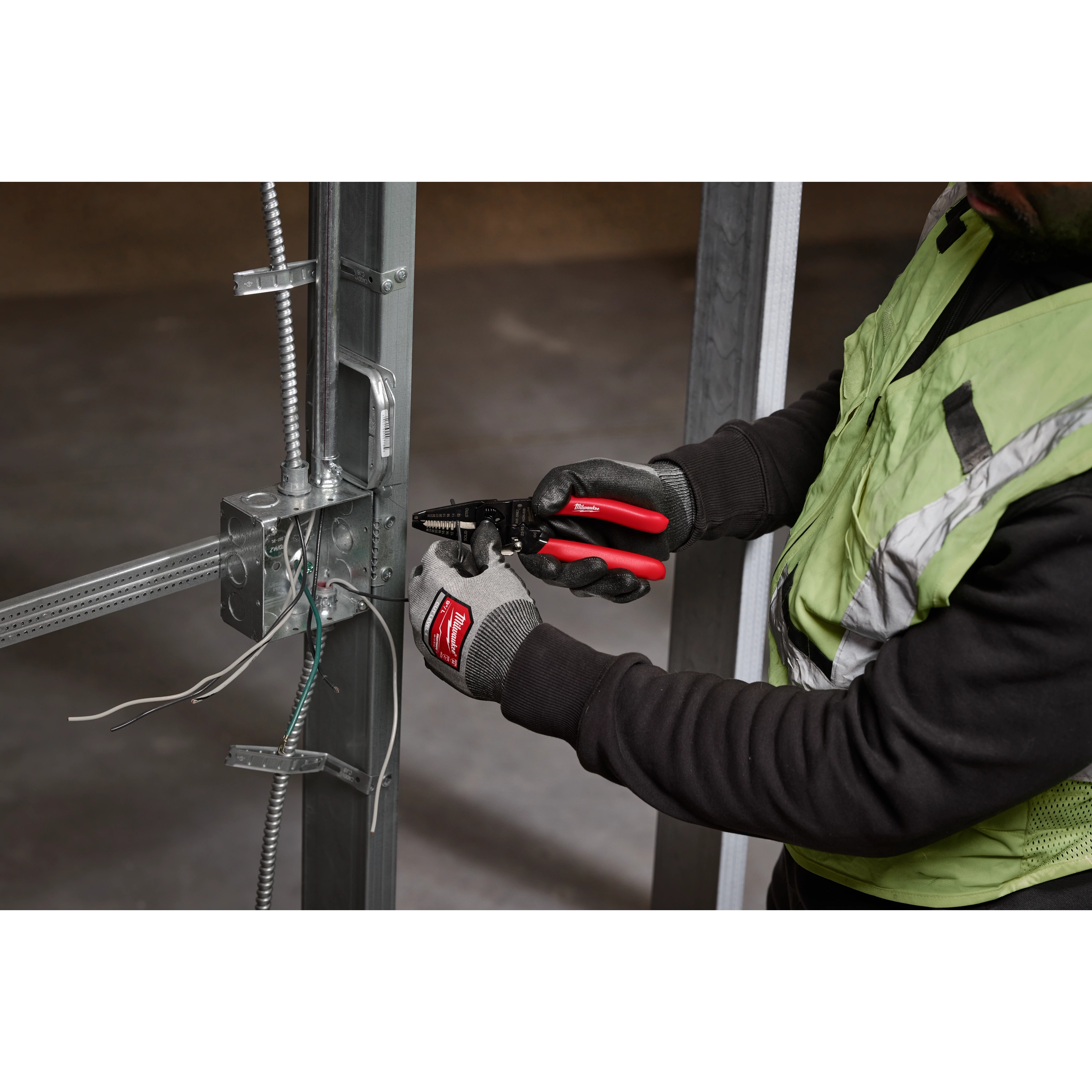 A person in work gloves uses a 10-28 AWG Multi-Purpose Dipped Grip Wire Stripper & Cutter with Reinforced Head to strip wires on a metal frame structure.