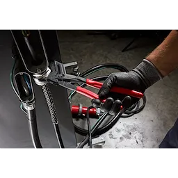 A person wearing black gloves uses a 10" Pliers Wrench to tighten a metal connector on industrial machinery. The tool has a black body and red handles, designed for gripping and turning purposes in mechanical applications.