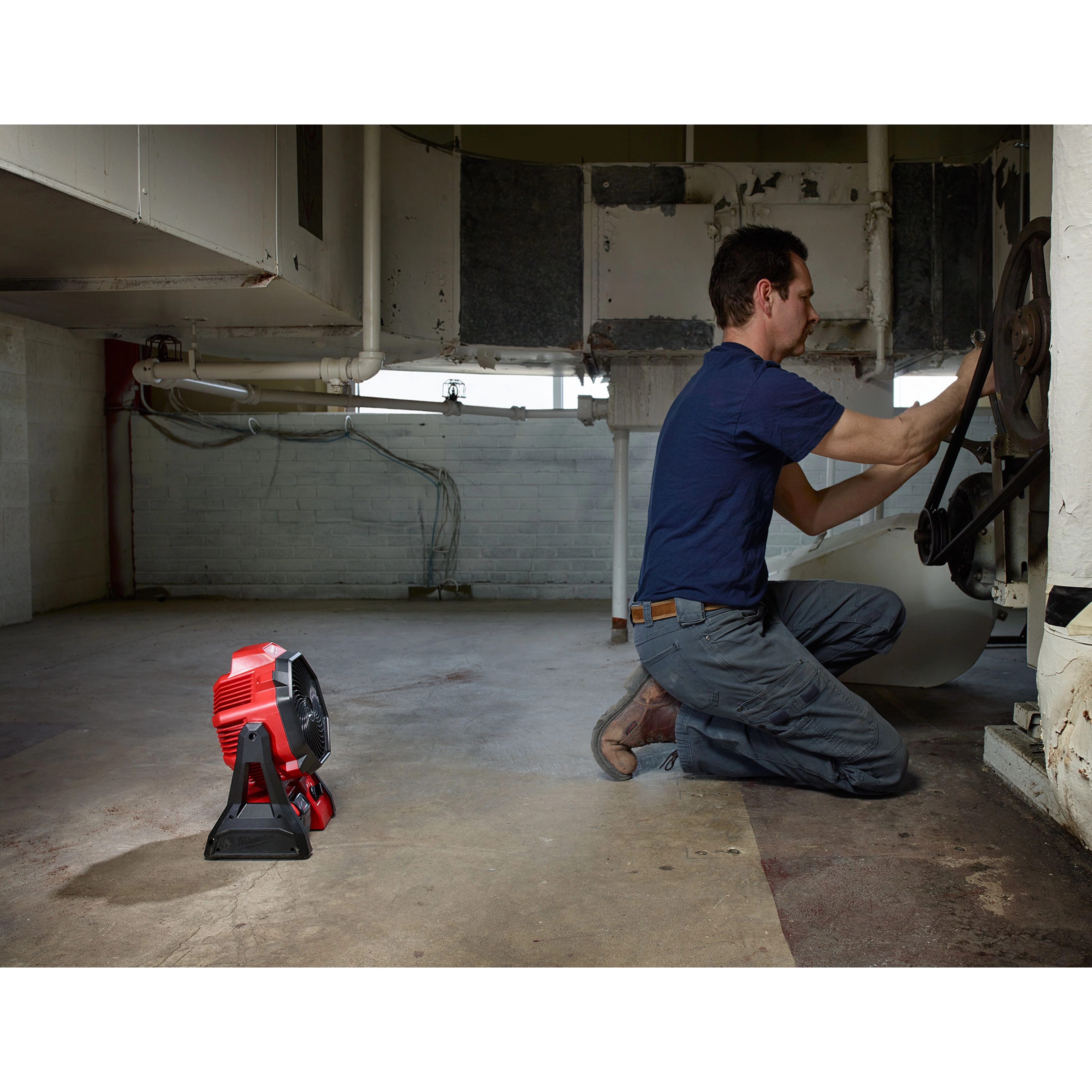 A worker kneels to repair machinery in a dimly-lit room with exposed pipes while the red and black M18 Jobsite Fan stands nearby.