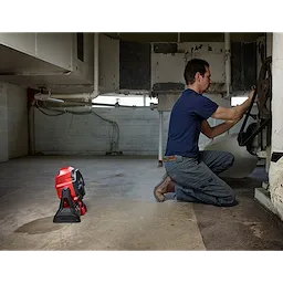 A worker kneels to repair machinery in a dimly-lit room with exposed pipes while the red and black M18 Jobsite Fan stands nearby.