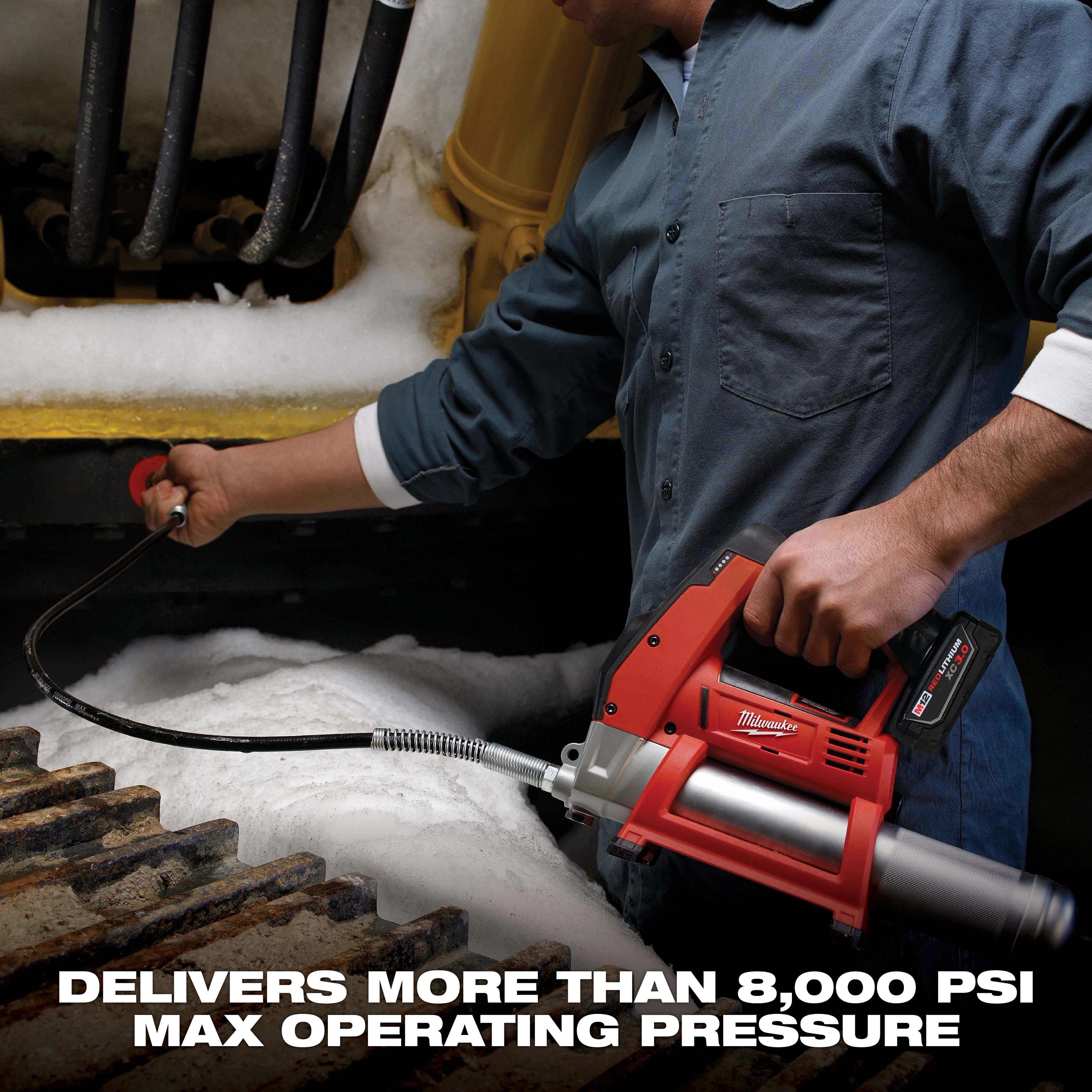 A person operates a Milwaukee cordless grease gun under snowy industrial equipment. The grease gun, held in the person's right hand, is connected to a hose. Text at the bottom reads, "Delivers more than 8,000 PSI max operating pressure."
