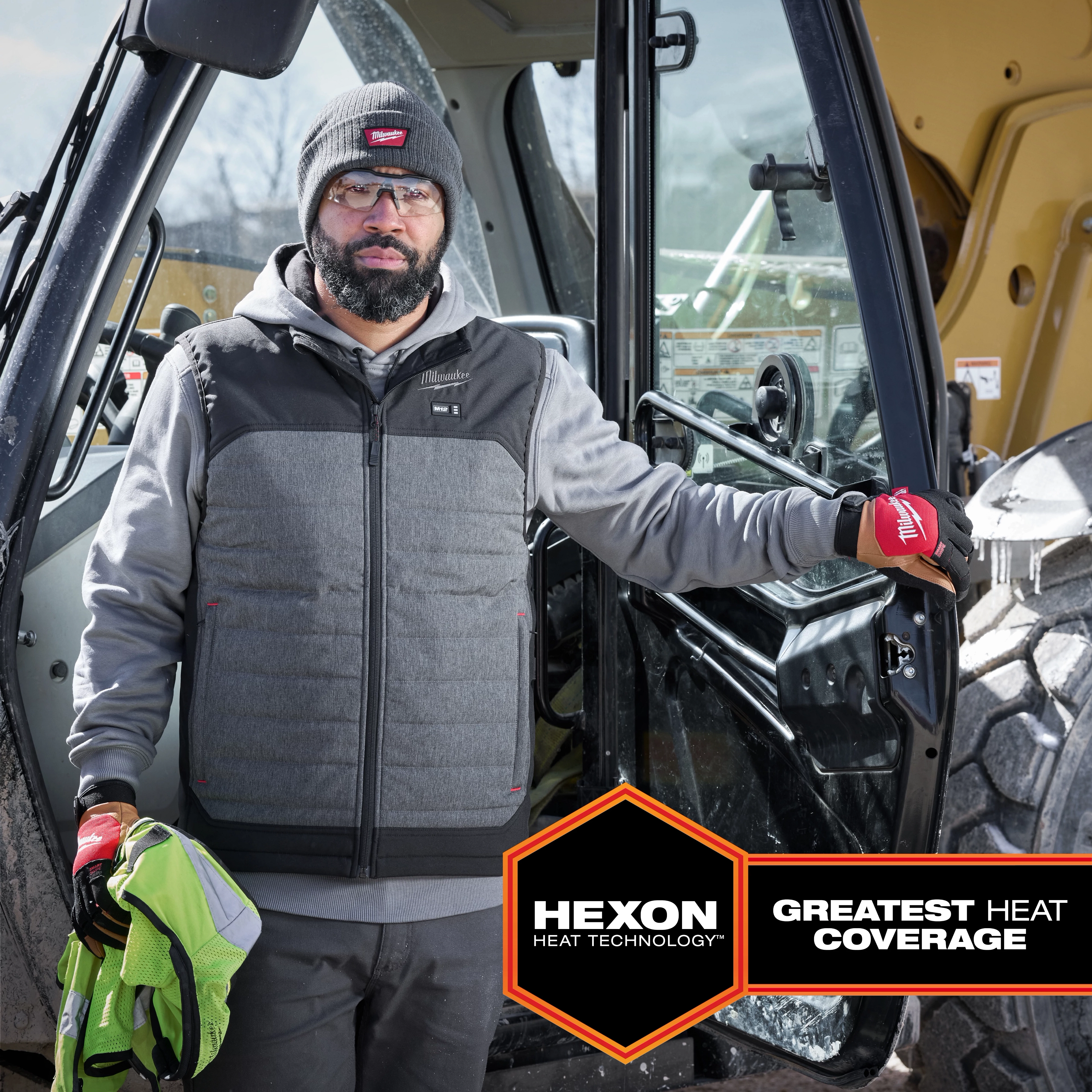 Man wearing M12 Heated AXIS Vest, standing beside construction machinery. Vest has Hexon heat technology offering greatest heat coverage.