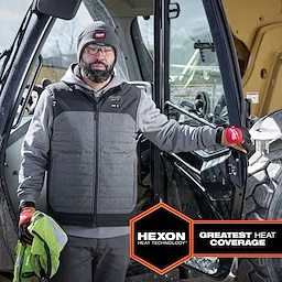 Man wearing M12 Heated AXIS Vest, standing beside construction machinery. Vest has Hexon heat technology offering greatest heat coverage.