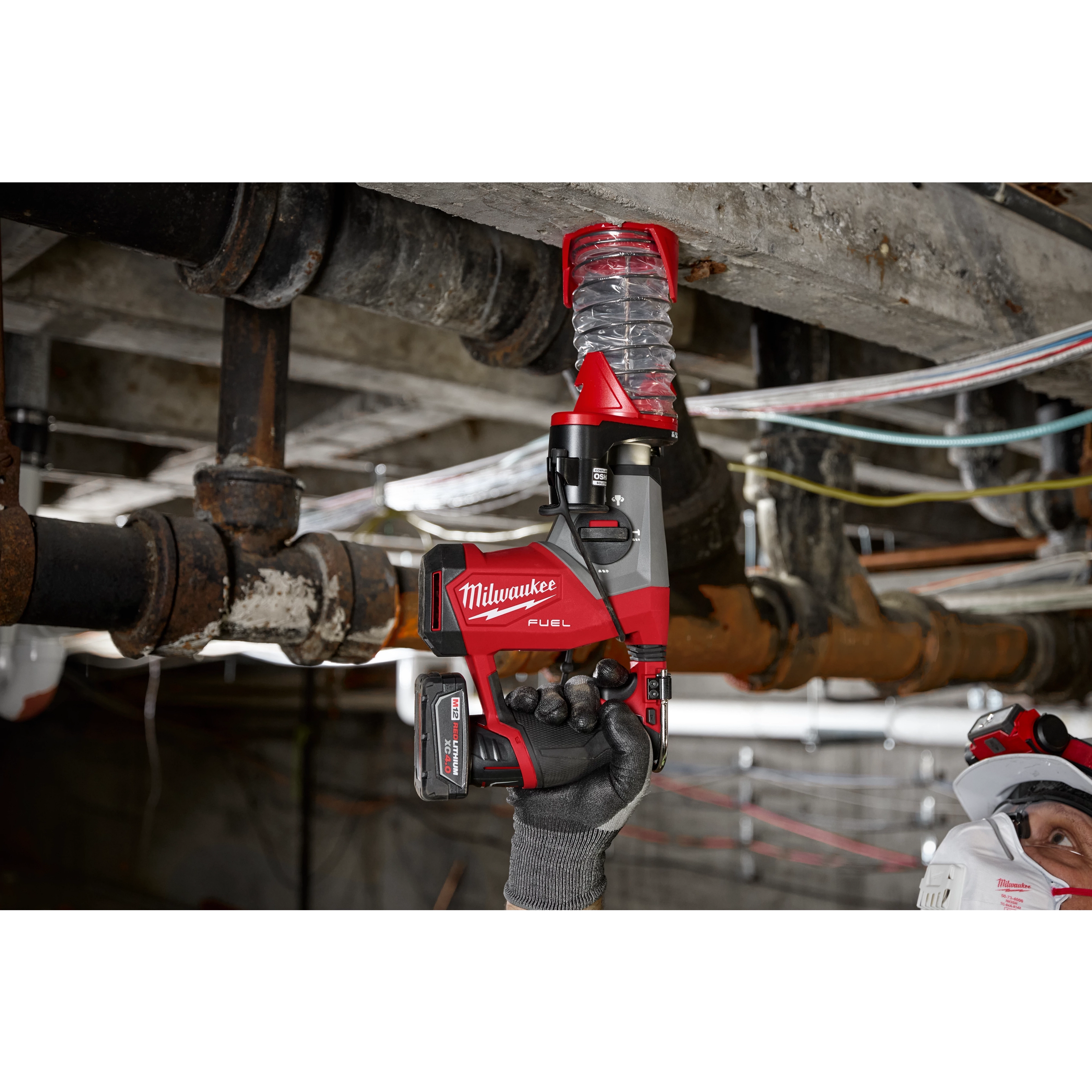 A person using the M12 FUEL™ 5/8” SDS Plus Rotary Hammer Kit is drilling into a concrete ceiling. The red and black tool is held upwards, and the person is wearing safety gear, including a helmet and mask, in an industrial setting with exposed pipes and cables.