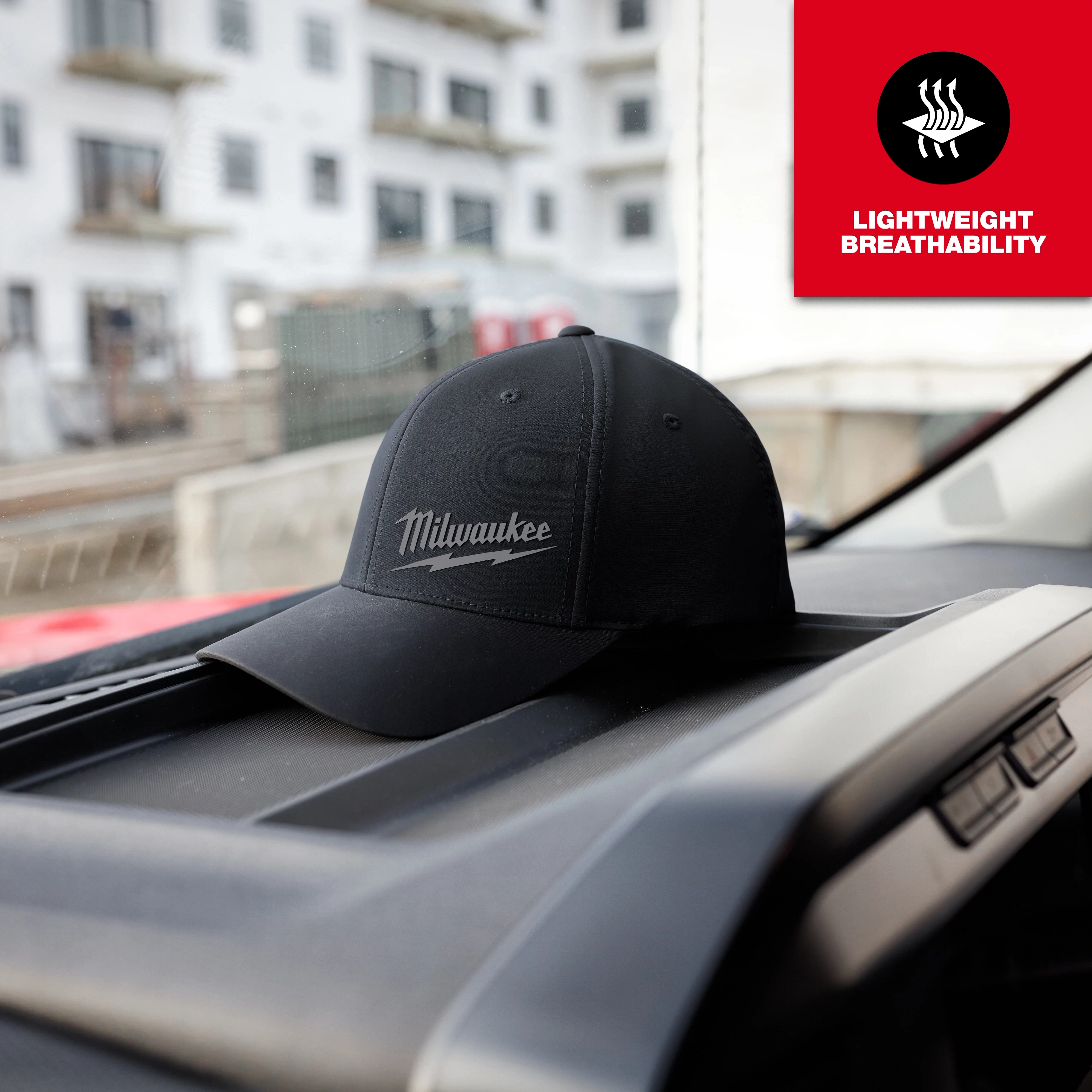 Black WORKSKIN Performance Fitted Hat with "Milwaukee" logo on dashboard. Red icon reads "Lightweight Breathability." Building visible outside.