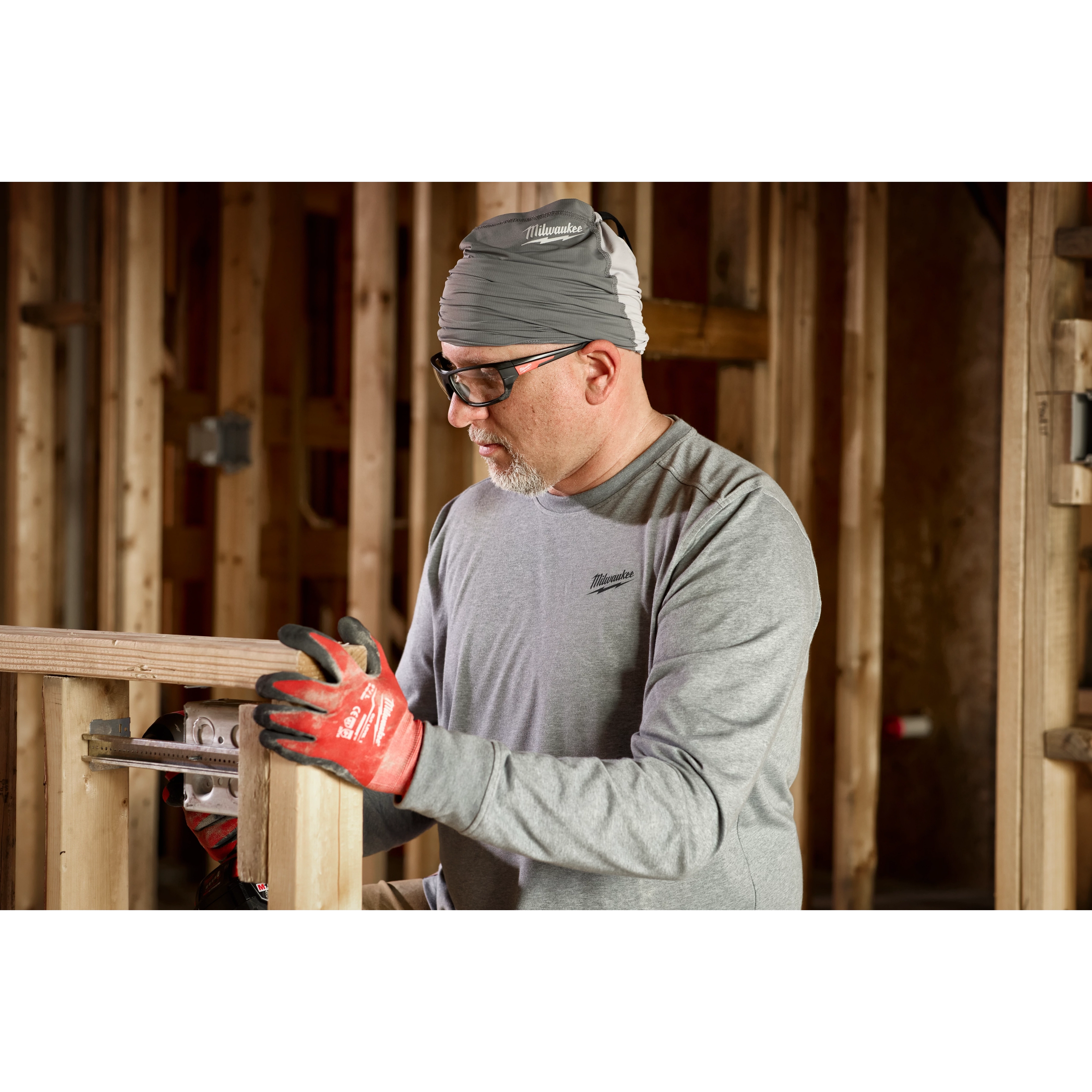 Person working indoors wearing a long-sleeve shirt, red gloves, and gray WORKSKIN Performance Neck Gaiter, handling a tool.