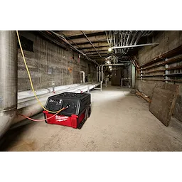 A ROLL-ON™ 7200W/3600W 6.0kWh Power Supply is positioned in an industrial basement with exposed pipes and wiring along concrete walls. The red and black unit has wheels for mobility and its cables are connected, suggesting active use. The product is designed for heavy-duty, portable power in demanding environments.