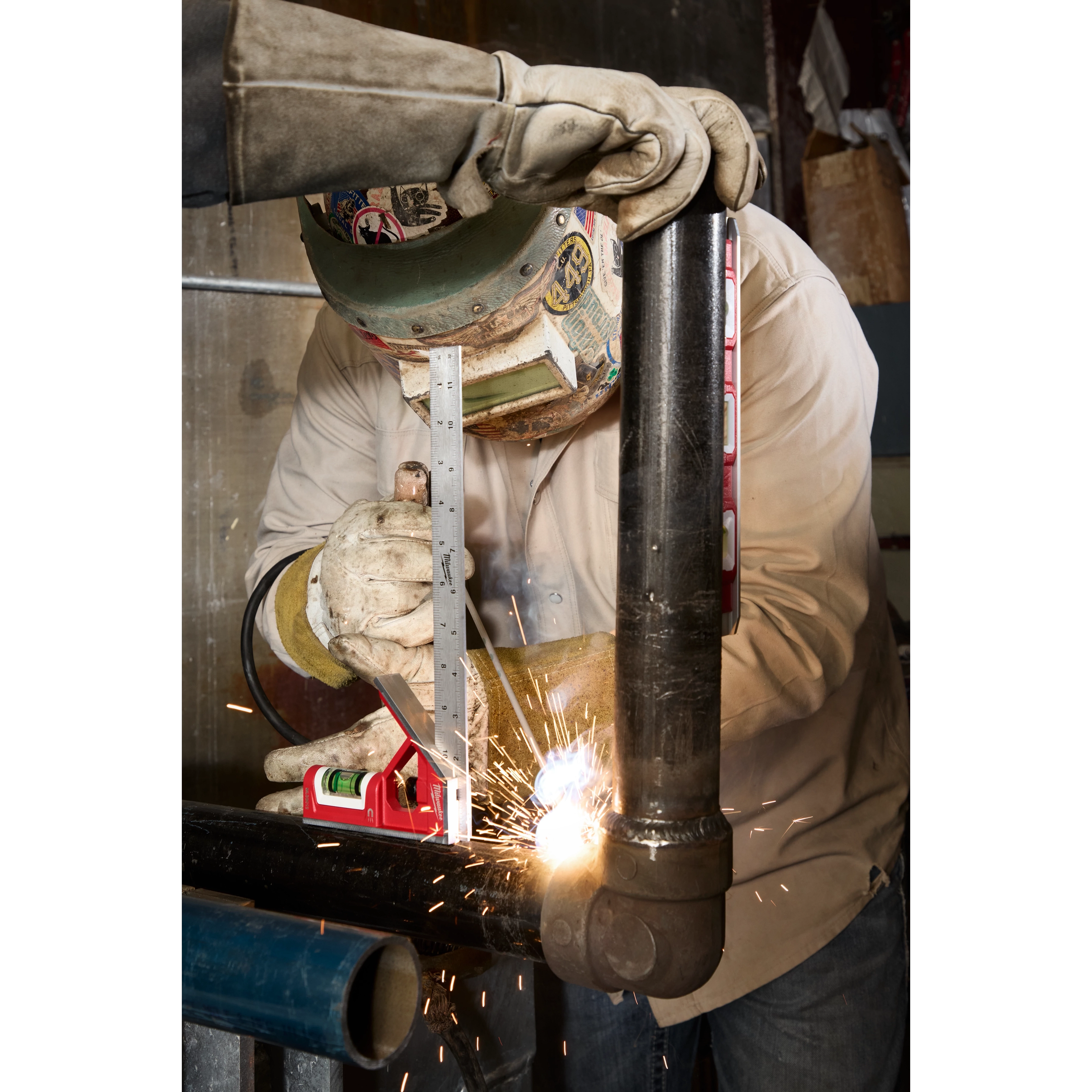 Welder using a 12" Magnetic Combination Square to measure a metal pipe with a spirit level attached. Welding sparks visible.