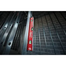 REDSTICK Level, a red level tool with bubble vials, is vertically placed against a metal frame in a construction site.