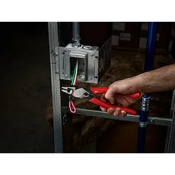 A person uses 9" High Leverage Lineman's Pliers With Crimper to grasp wires near an electrical box on a metal frame.