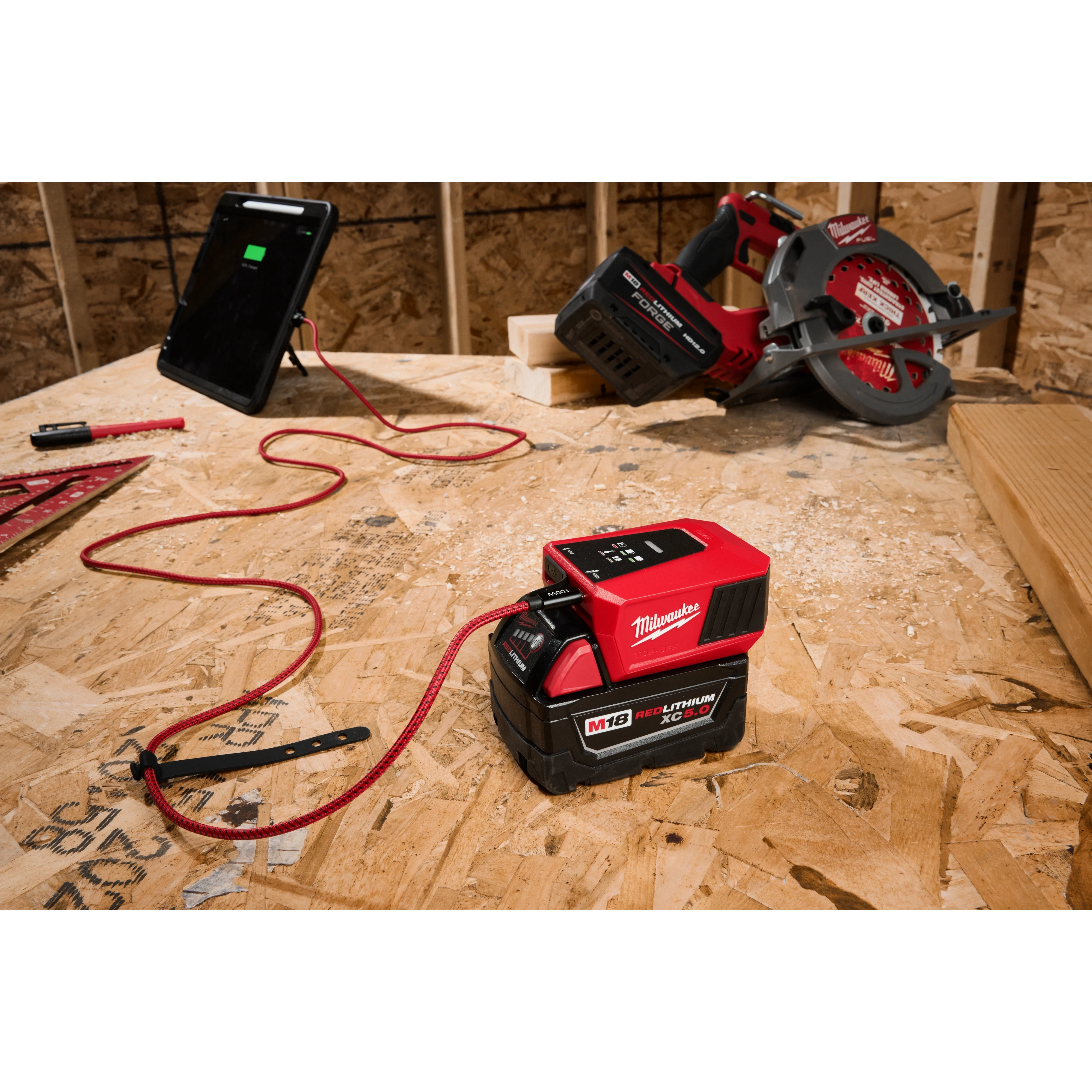 The M18™ TOP-OFF™ Charger & Power Supply is on a wooden workbench, connected to a tablet via a red charging cable, with a power tool and other construction equipment visible in the background.