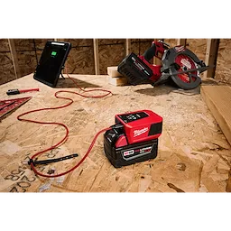 The M18™ TOP-OFF™ Charger & Power Supply is on a wooden workbench, connected to a tablet via a red charging cable, with a power tool and other construction equipment visible in the background.