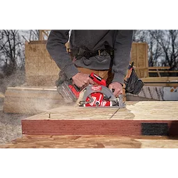 A worker uses an M18 FUEL™ 7-1/4” Circular Saw to cut through a piece of wood on an outdoor construction site. Sawdust is visible coming from the cutting area, and the worker is wearing a gray sweatshirt and brown work pants. Various construction materials are in the background.