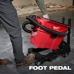 A person operates the NEXUS™ Wet/Dry Vacuum Cart, a red Milwaukee-branded vacuum with a foot pedal and hose attachment, positioned on a wheeled cart in a work area with tools and debris. The text "FOOT PEDAL" is displayed at the bottom.