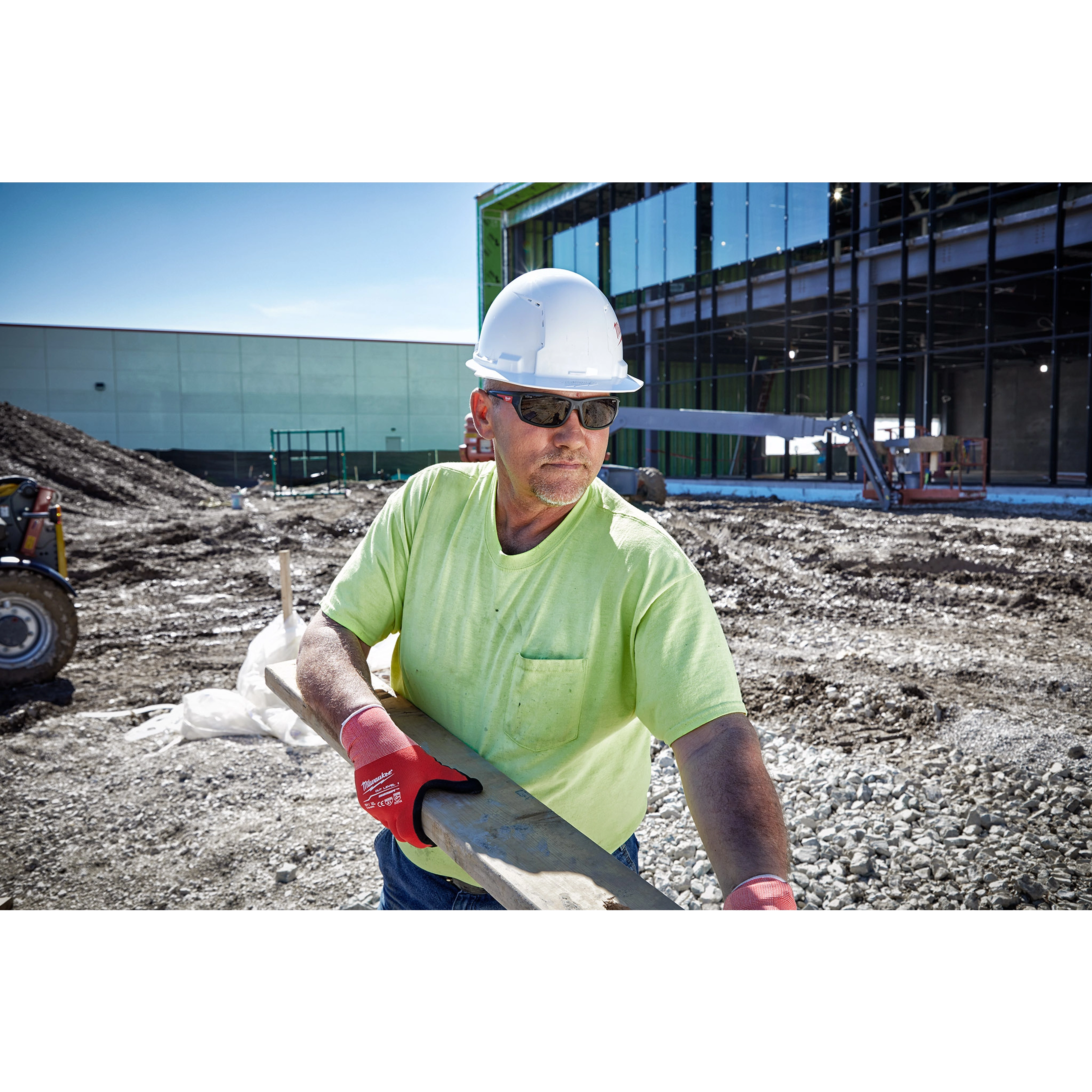 A construction worker wearing Safety Glasses - Tinted Anti-Scratch Lenses, a white hard hat, and red gloves holding a wooden plank.