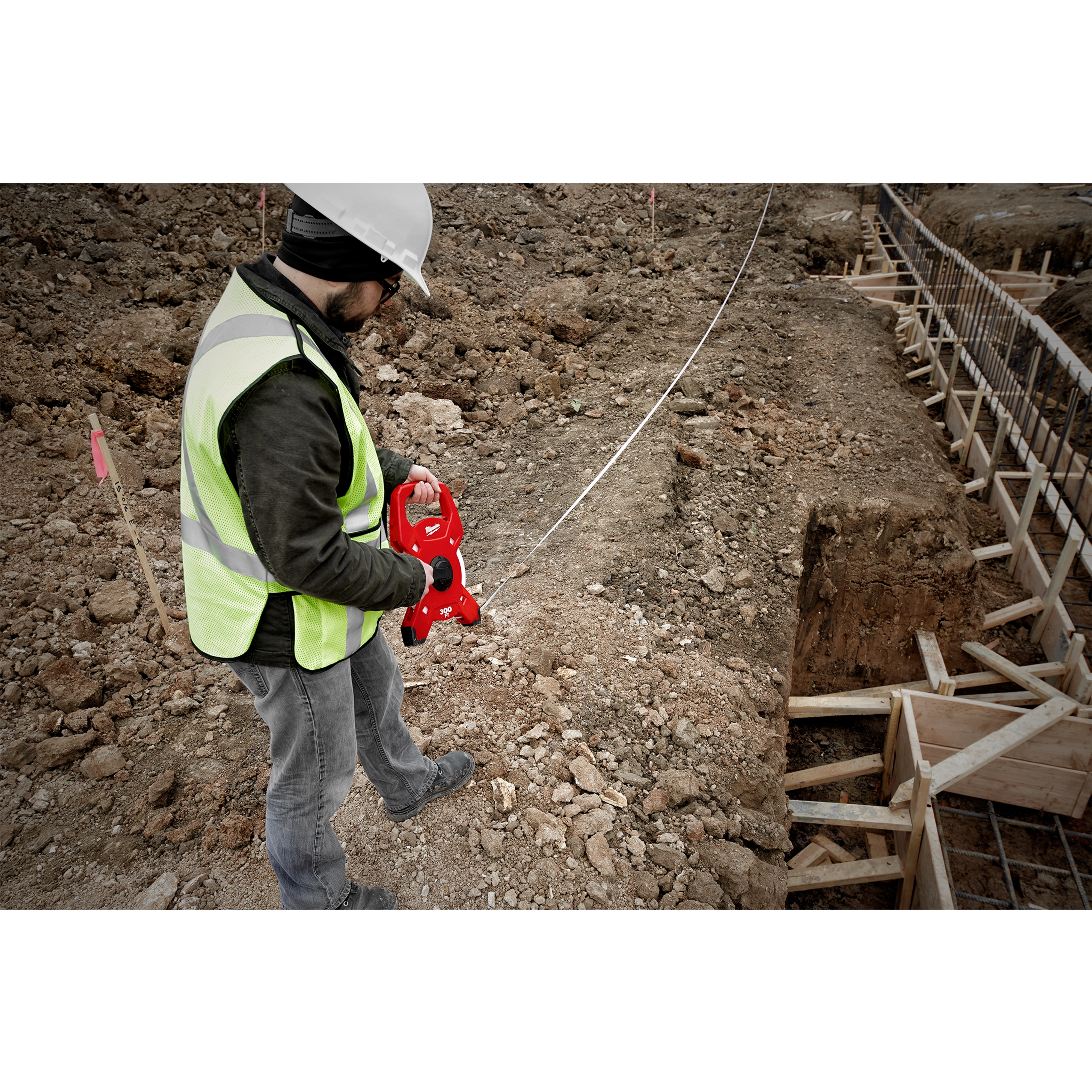 A construction worker uses a Milwaukee 300 Foot Fiberglass Long Tape while measuring on a rugged construction site with soil and wooden frames.