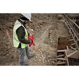 A construction worker uses a Milwaukee 300 Foot Fiberglass Long Tape while measuring on a rugged construction site with soil and wooden frames.