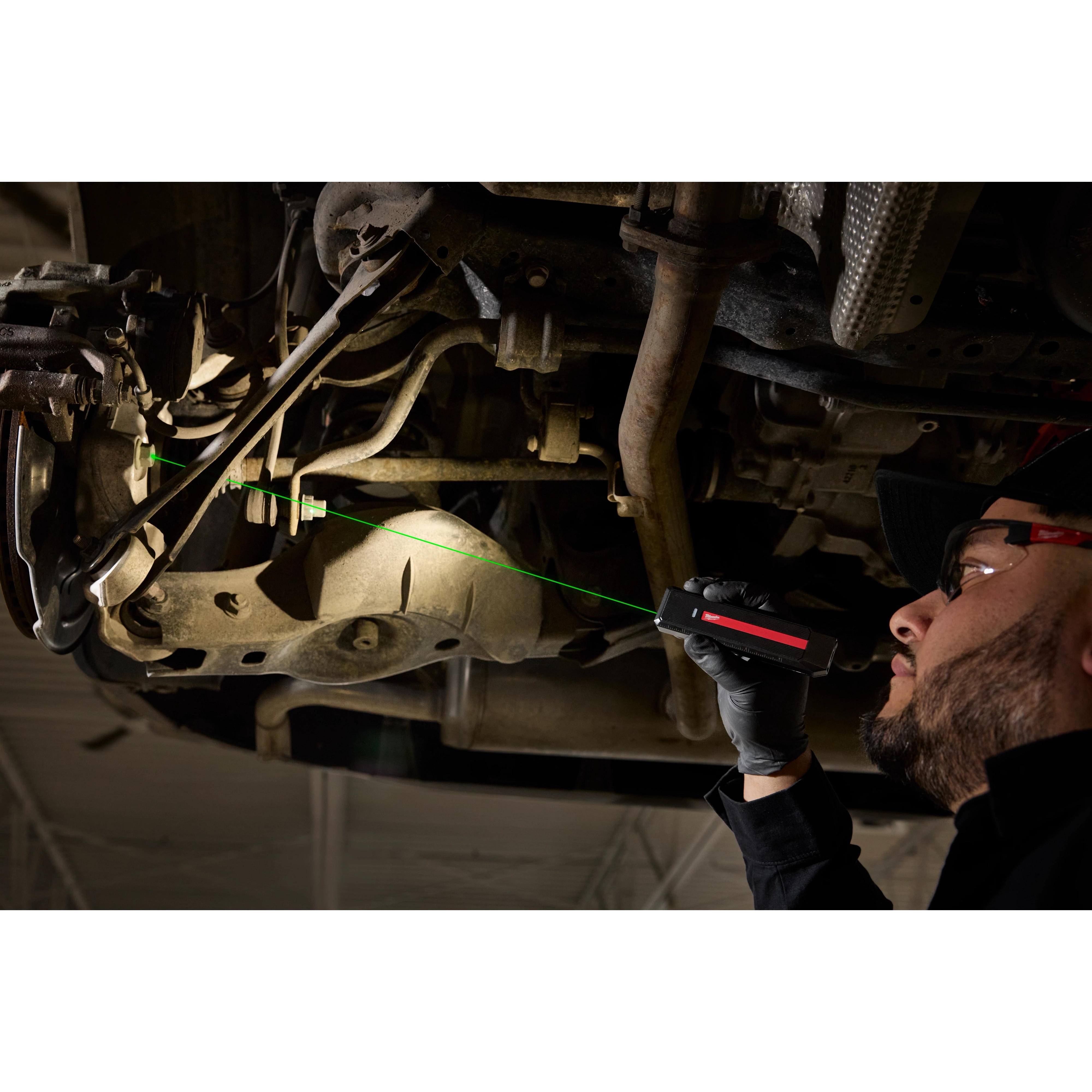 A person using a Milwaukee Rechargeable Flat Flashlight with Green Laser to inspect the underside of a vehicle in a workshop.