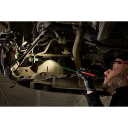 A person using a Milwaukee Rechargeable Flat Flashlight with Green Laser to inspect the underside of a vehicle in a workshop.