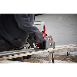 Person using the M18 FUEL 6 1/2" Plunge Track Saw to cut a wooden board outdoors on a portable workbench.