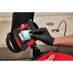 A gloved hand interacts with the touchscreen of an M12™ Wireless Monitor, which is mounted on a red device with the Milwaukee logo. The screen displays several icons and a video feed. The background features a concrete surface with a few holes.