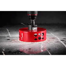A close-up image of a Milwaukee® 4-1/2" DIAMOND MAX™ Hole Saw, shown cutting through a smooth, reflective surface. The bright red hole saw is attached to a drill, and there is dust and fragments around the cutting area.