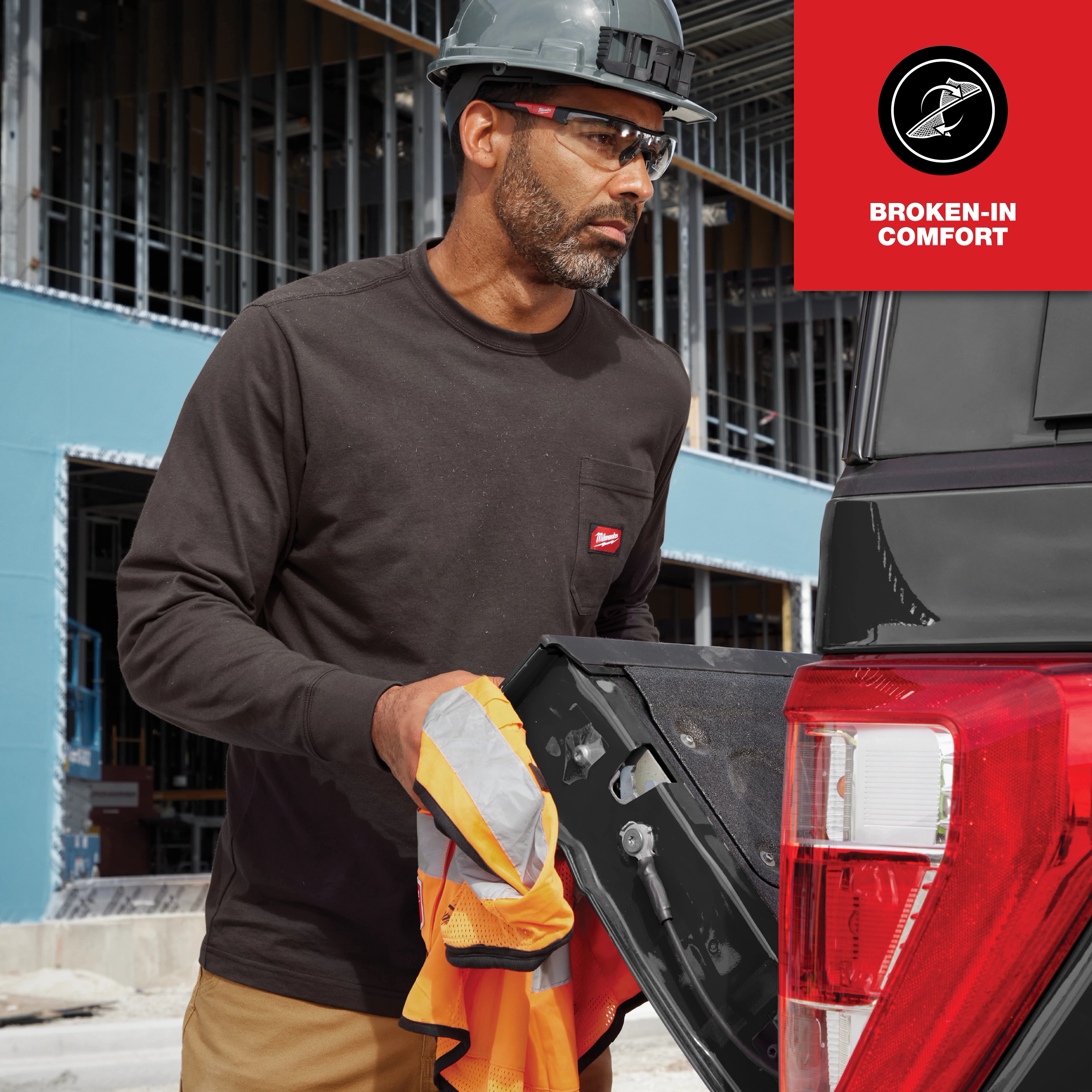 A person wearing a long-sleeve GRIDIRON Pocket T-Shirt in a construction setting, standing near an open vehicle tailgate and holding a safety vest.