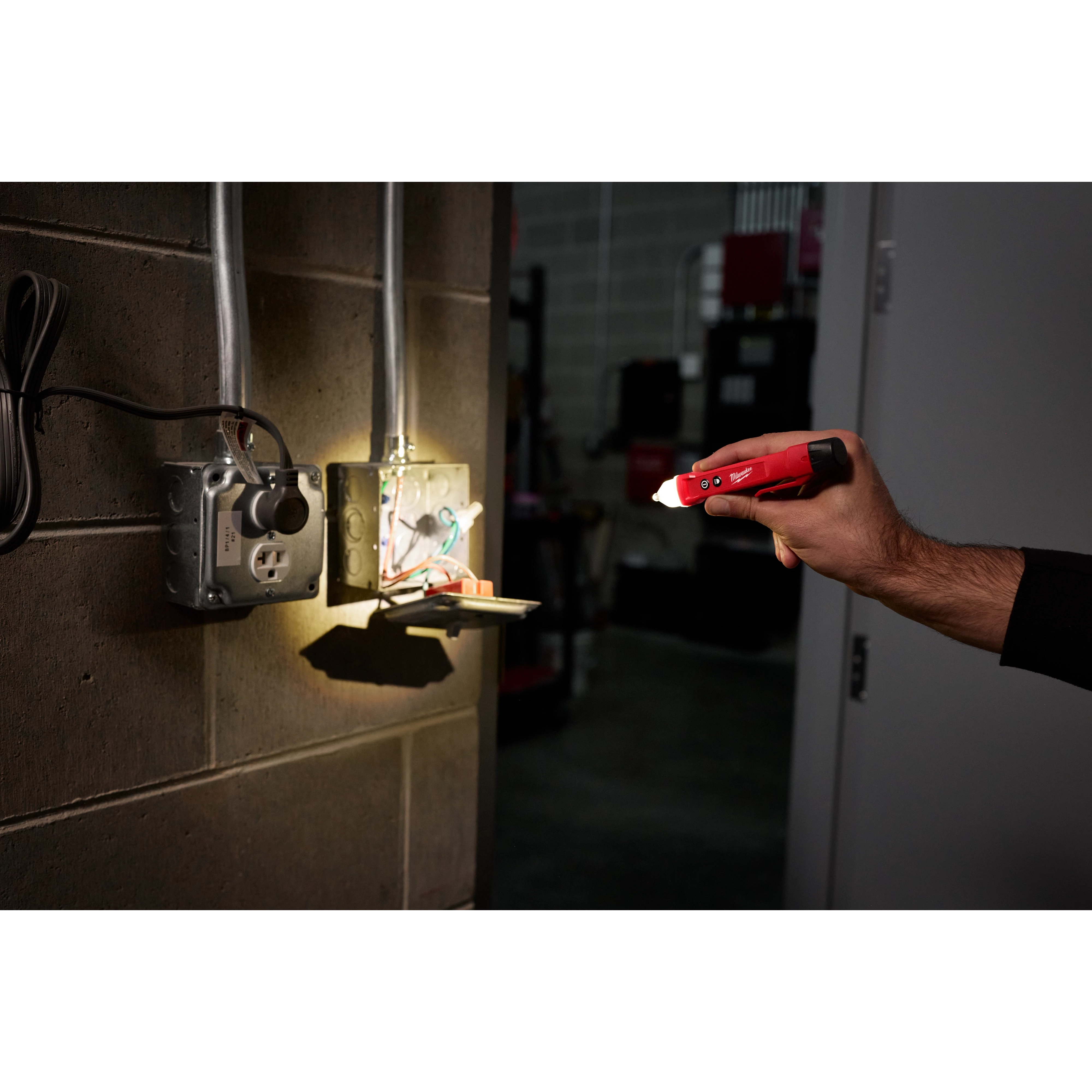 A person uses a Voltage Detector w/ LED to test an open electrical outlet box in a dimly lit room. The detector's LED light is illuminated, highlighting the wiring inside the electrical box.