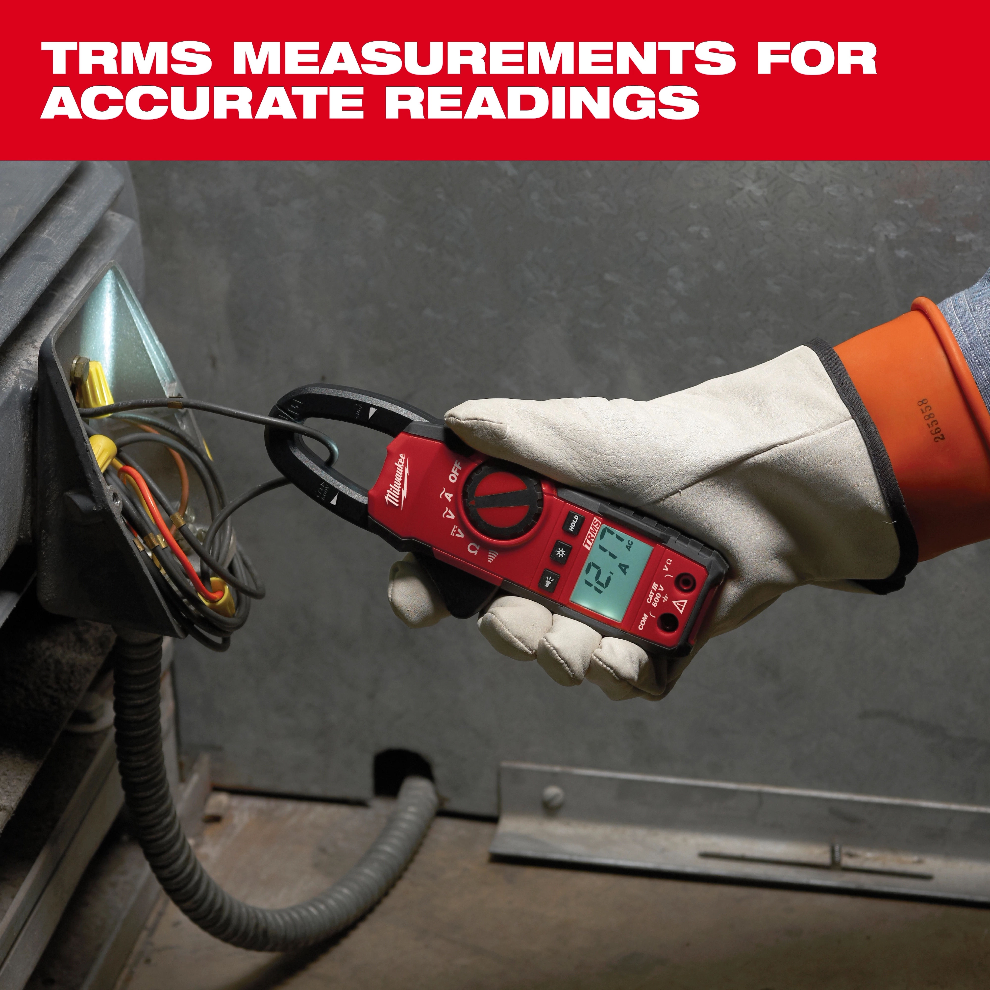 A gloved hand uses a Milwaukee electrical meter to measure voltage on a set of wires in an industrial panel. The meter shows a reading of 12.1 volts. The red device has a digital display and clamp for securing around wires. Text above reads: "TRMS Measurements for Accurate Readings."