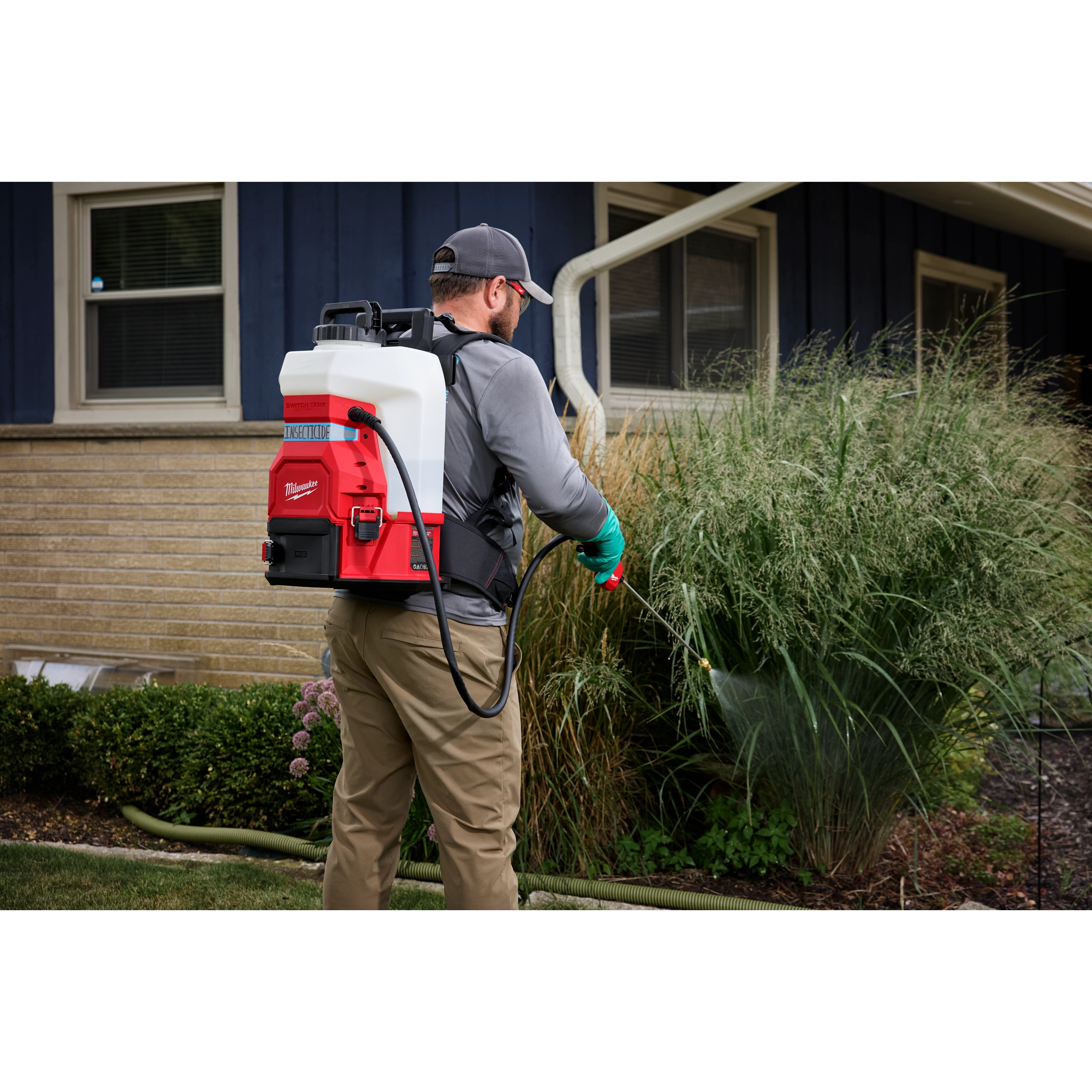 A person wearing a gray cap and gloves uses a red and white Milwaukee backpack sprayer to water plants beside a house. The person stands on a lawn next to a beige and blue house with two windows.