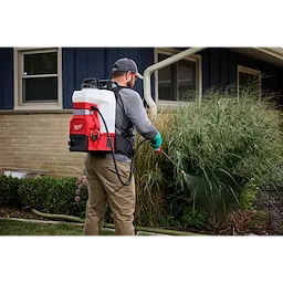 A person wearing a gray cap and gloves uses a red and white Milwaukee backpack sprayer to water plants beside a house. The person stands on a lawn next to a beige and blue house with two windows.