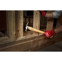 Person using a 19oz Milled Face Hickory Hammer to drive a nail into a wooden frame, wearing red work gloves.
