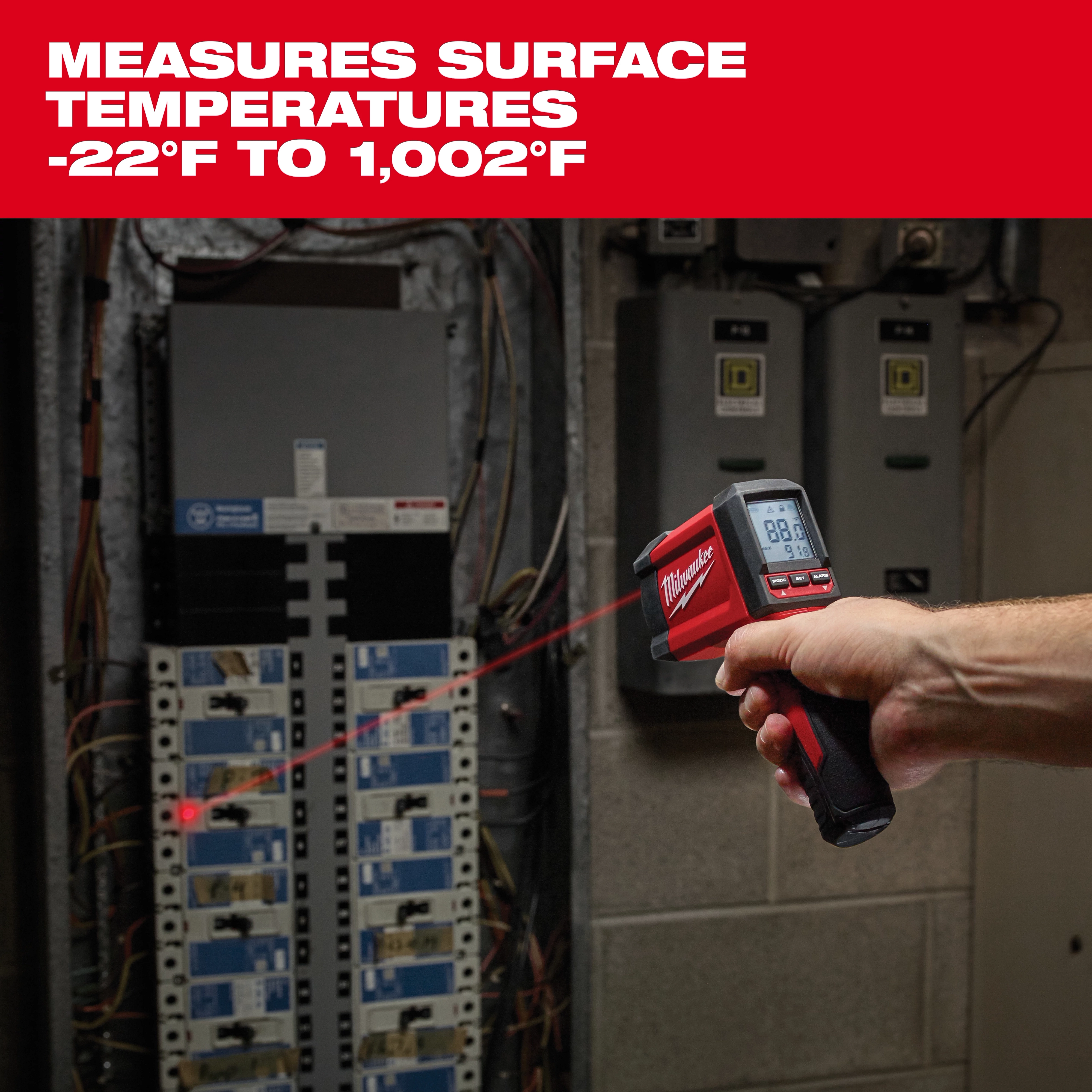 A hand-held Milwaukee infrared thermometer measures surface temperatures ranging from -22°F to 1,002°F. The device displays a reading on its digital screen and points a laser at electrical panels in an industrial setting. The product's logo appears prominently on its body.