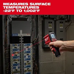 A hand-held Milwaukee infrared thermometer measures surface temperatures ranging from -22°F to 1,002°F. The device displays a reading on its digital screen and points a laser at electrical panels in an industrial setting. The product's logo appears prominently on its body.