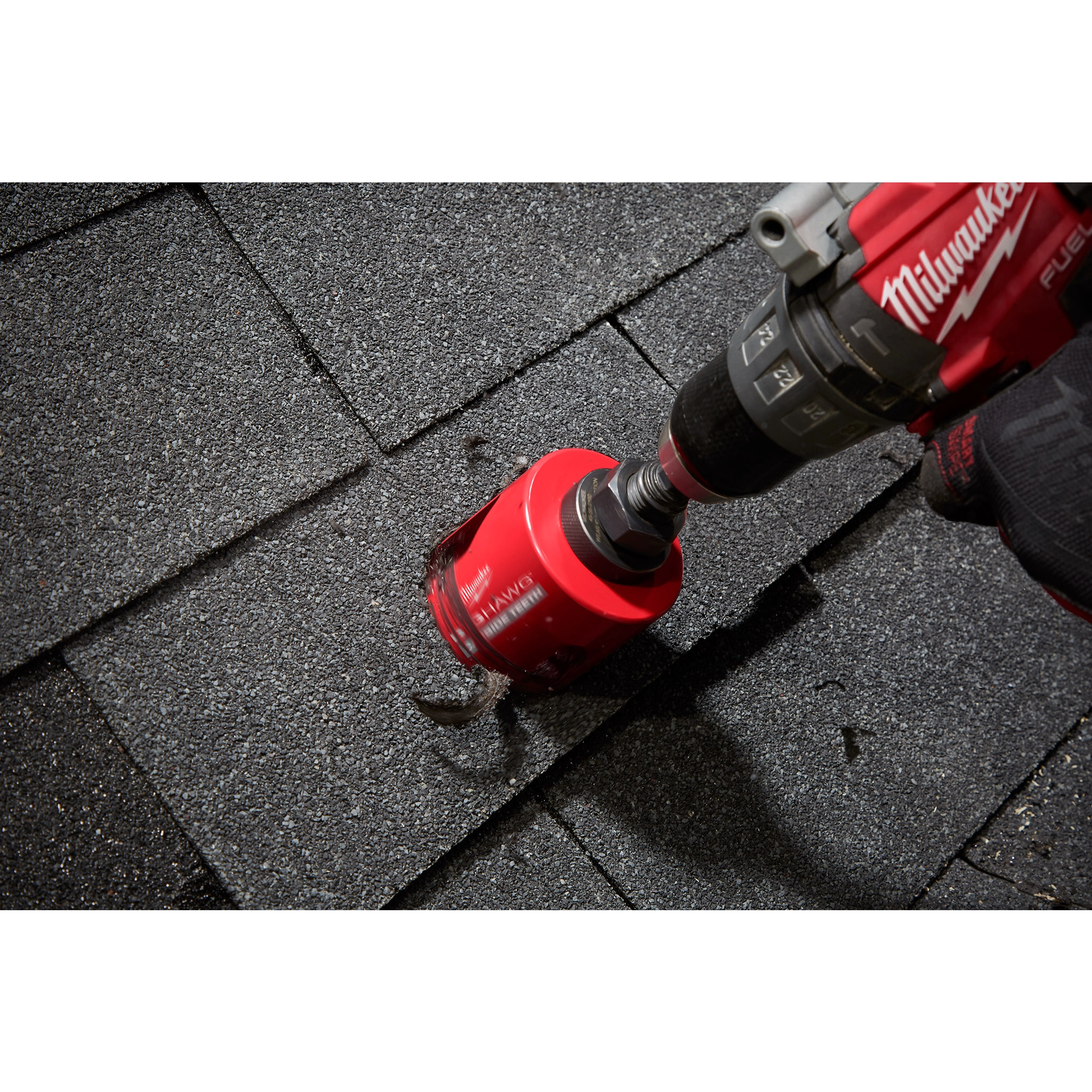 A red 2-9/16" BIG HAWG™ with Carbide Teeth is attached to a power drill, cutting into black asphalt roofing shingles. The tool appears to be in motion, creating a circular hole in the roofing material. Only hands operating the drill and the working area are visible.
