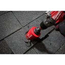 A red 2-9/16" BIG HAWG™ with Carbide Teeth is attached to a power drill, cutting into black asphalt roofing shingles. The tool appears to be in motion, creating a circular hole in the roofing material. Only hands operating the drill and the working area are visible.