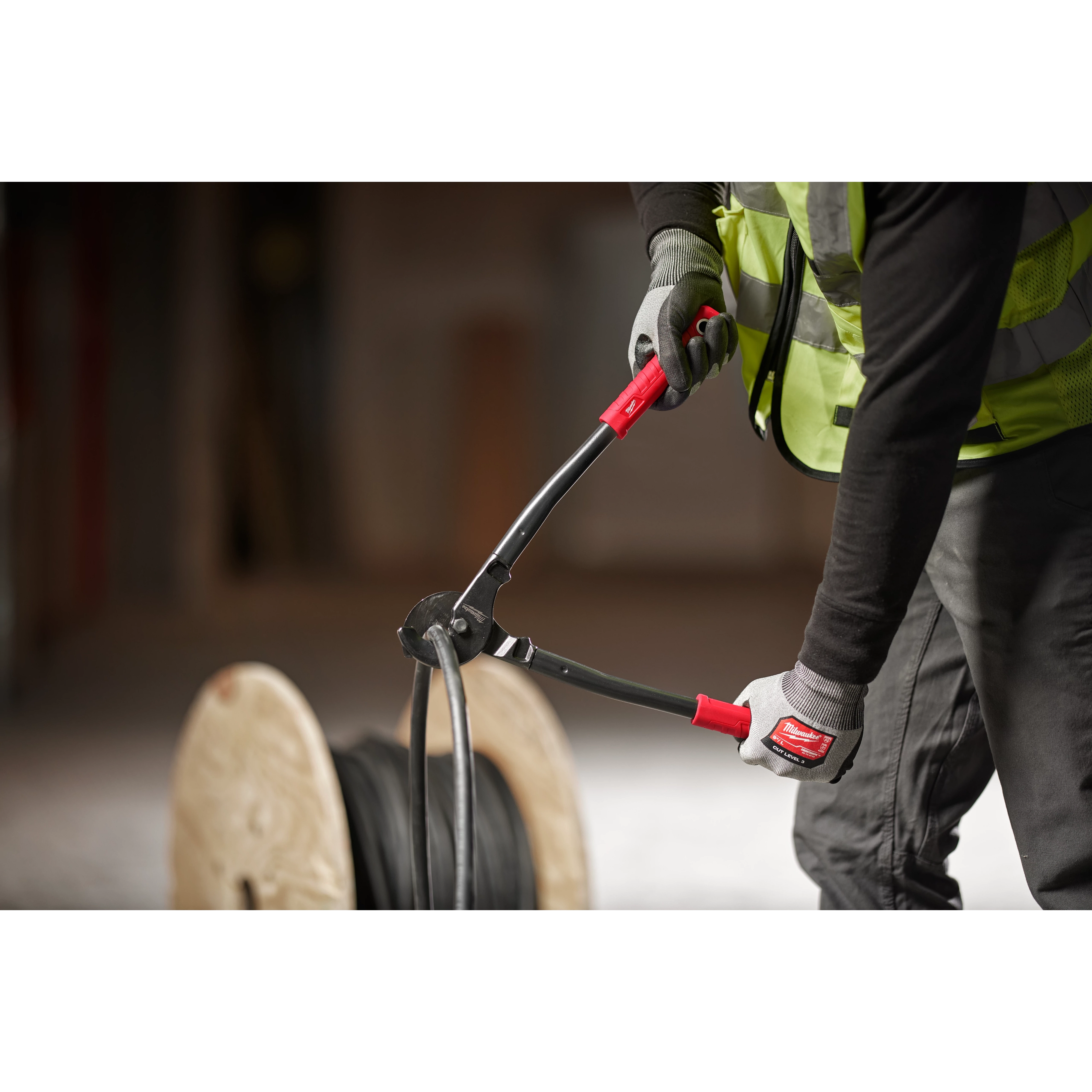 Person using the 17" Utility Cable Cutter to cut thick black cables, wearing a high-visibility vest and gloves, with a cable spool nearby.