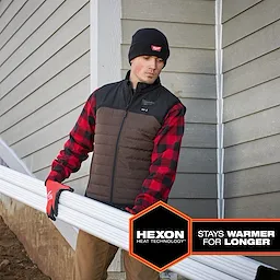 Person wearing a black and brown M12 Heated AXIS Vest with red-and-black checkered sleeves, holding white pipes. Hexon Heat Technology logo.
