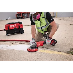A worker uses a Milwaukee 7"/9" Surface Grinding Dust Shroud attached to a grinder on a concrete surface. A vacuum dust collector is connected to a hose to efficiently manage debris.