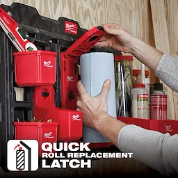 A person installing a blue paper roll onto a red PACKOUT Roll Holder attached to a wall with additional storage compartments.