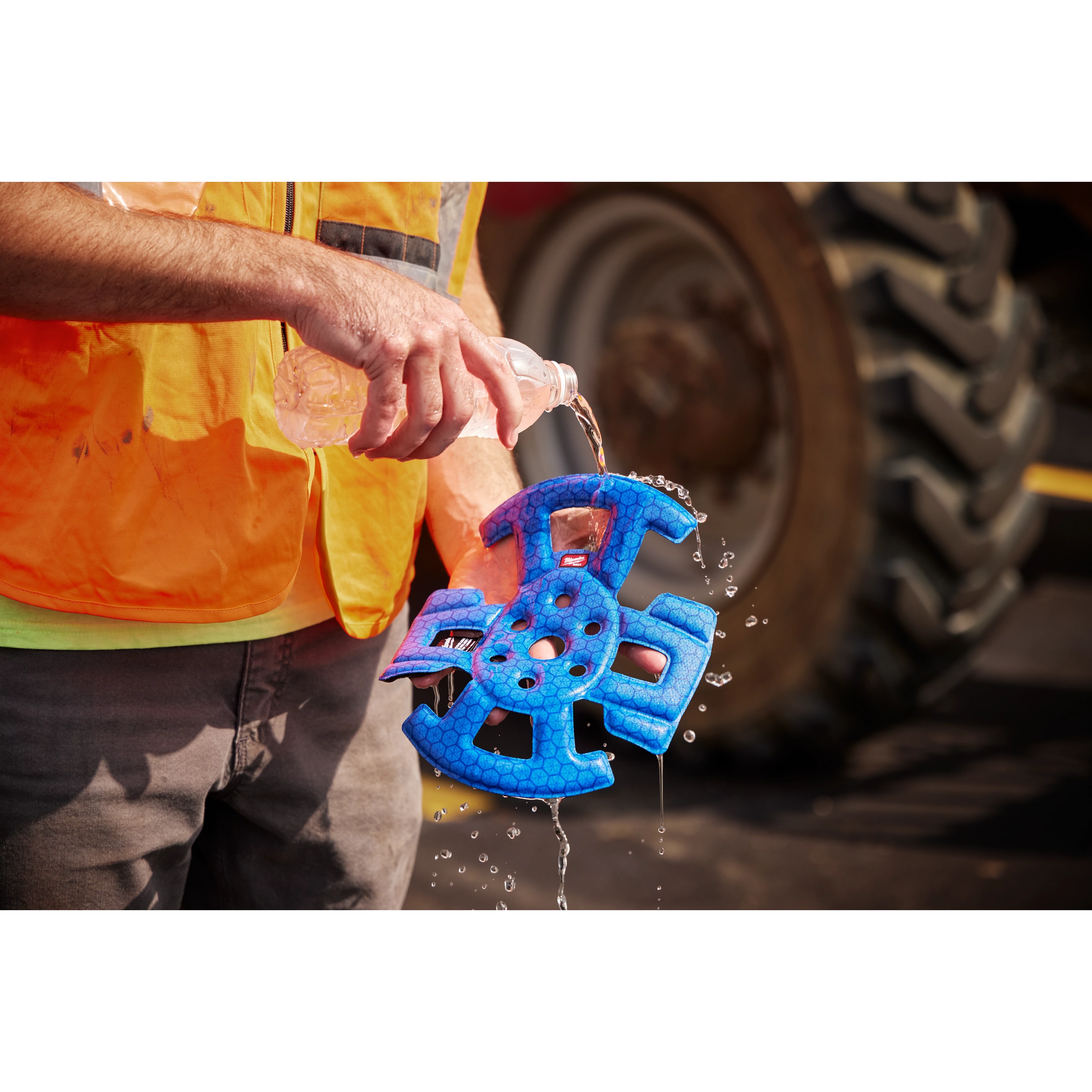 A construction worker pours water onto a blue BOLT™ Safety Helmet Cooling Liner. The worker is wearing an orange safety vest and grey pants, standing in front of heavy machinery. The liner is designed to keep helmets cool in hot conditions.