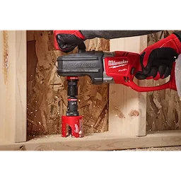 A person wearing red and black gloves uses a Hole Hawg drill with a 3/8" QUIK-LOK™ Arbor Set to drill into a wooden surface. The red drill is branded Milwaukee. Wood shavings are visible around the drilling area.