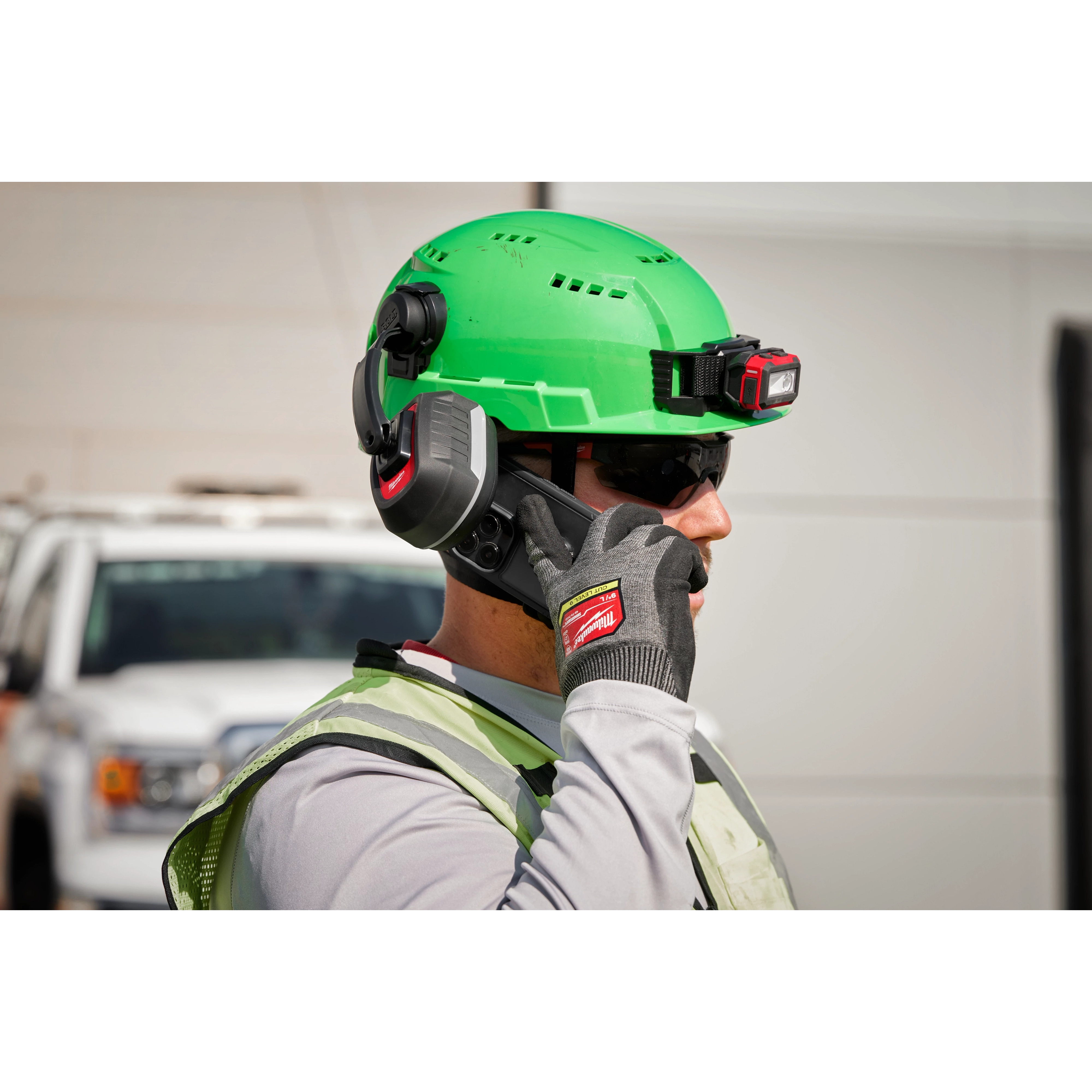 A worker wearing a green hard hat, safety vest, and BOLT 26dB Earmuffs stands in front of a white truck.