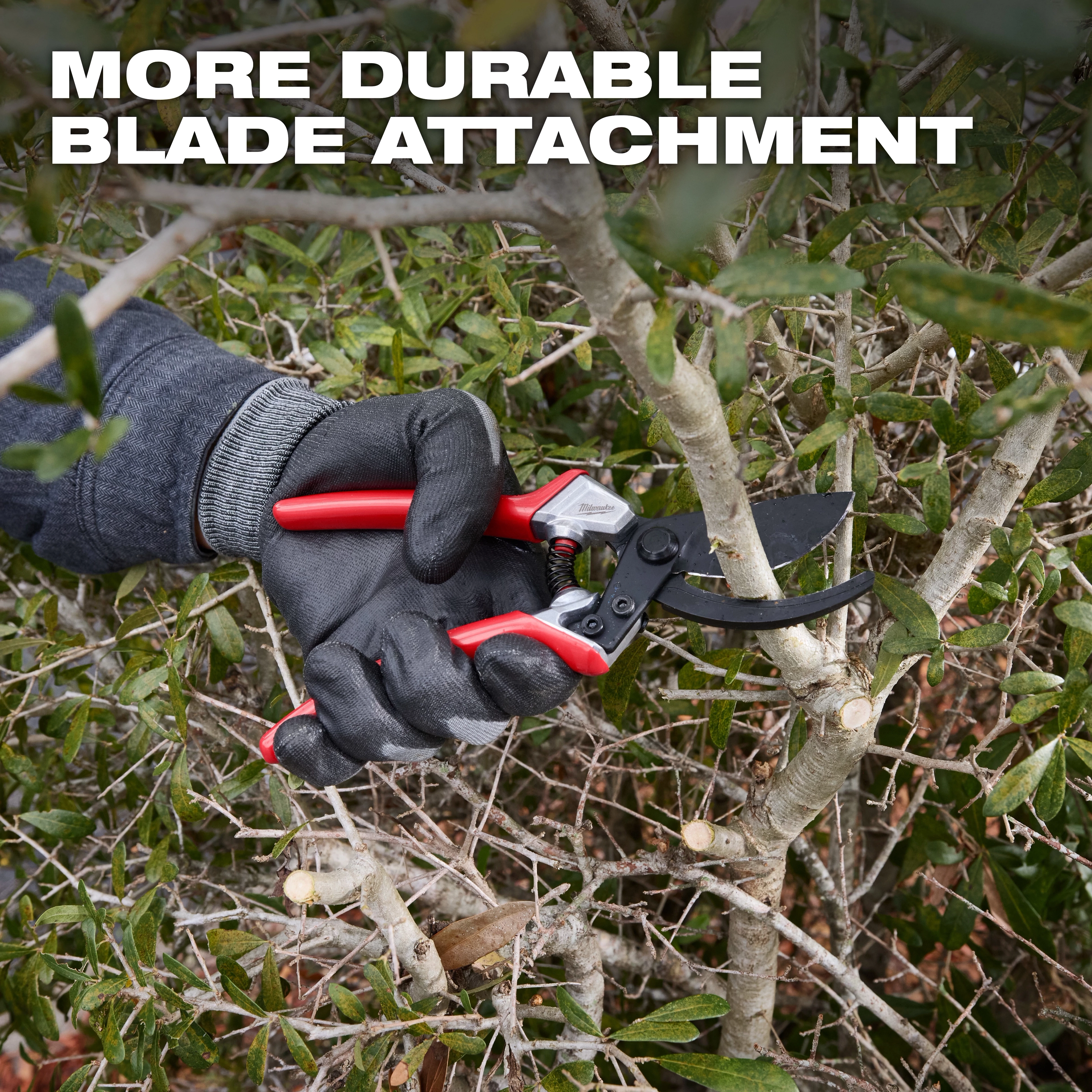 A gloved hand is using a 1" Pruner with a red handle to cut a branch from a bush. The text "MORE DURABLE BLADE ATTACHMENT" is displayed at the top of the image.