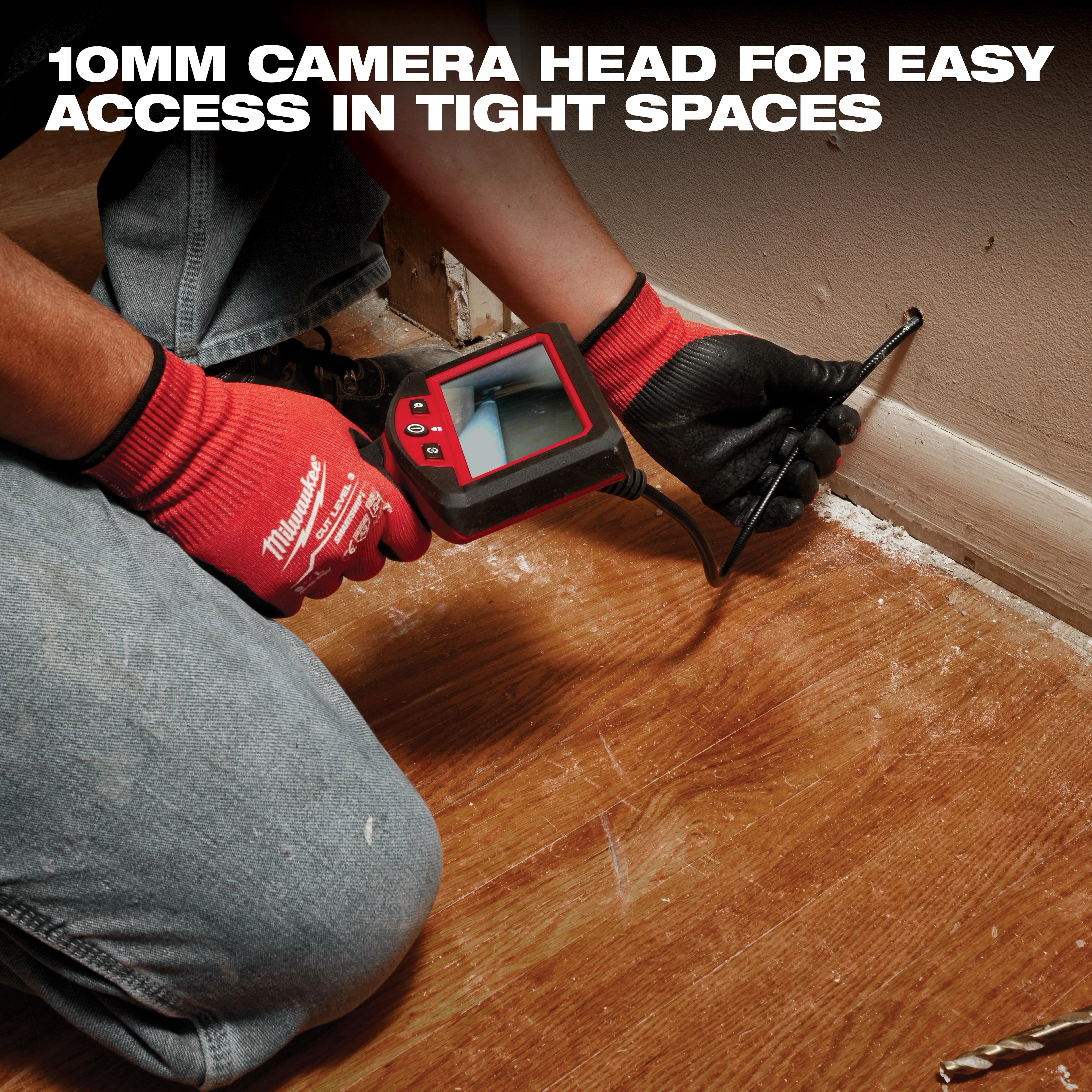A person in jeans and red gloves uses a Milwaukee inspection camera to view inside a tight space near a wooden floor and wall. The camera features a 10mm head for easy access in confined areas.
