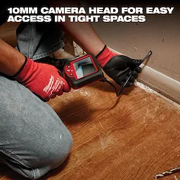 A person in jeans and red gloves uses a Milwaukee inspection camera to view inside a tight space near a wooden floor and wall. The camera features a 10mm head for easy access in confined areas.