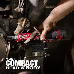 A person's hand uses the Milwaukee M12 FUEL™ 1/4" Ratchet to tighten a pipe fitting in a mechanical setup. The ratchet's compact head and body help in accessing tight spaces. Bold text reads "MOST COMPACT HEAD & BODY" in the image's bottom left corner.