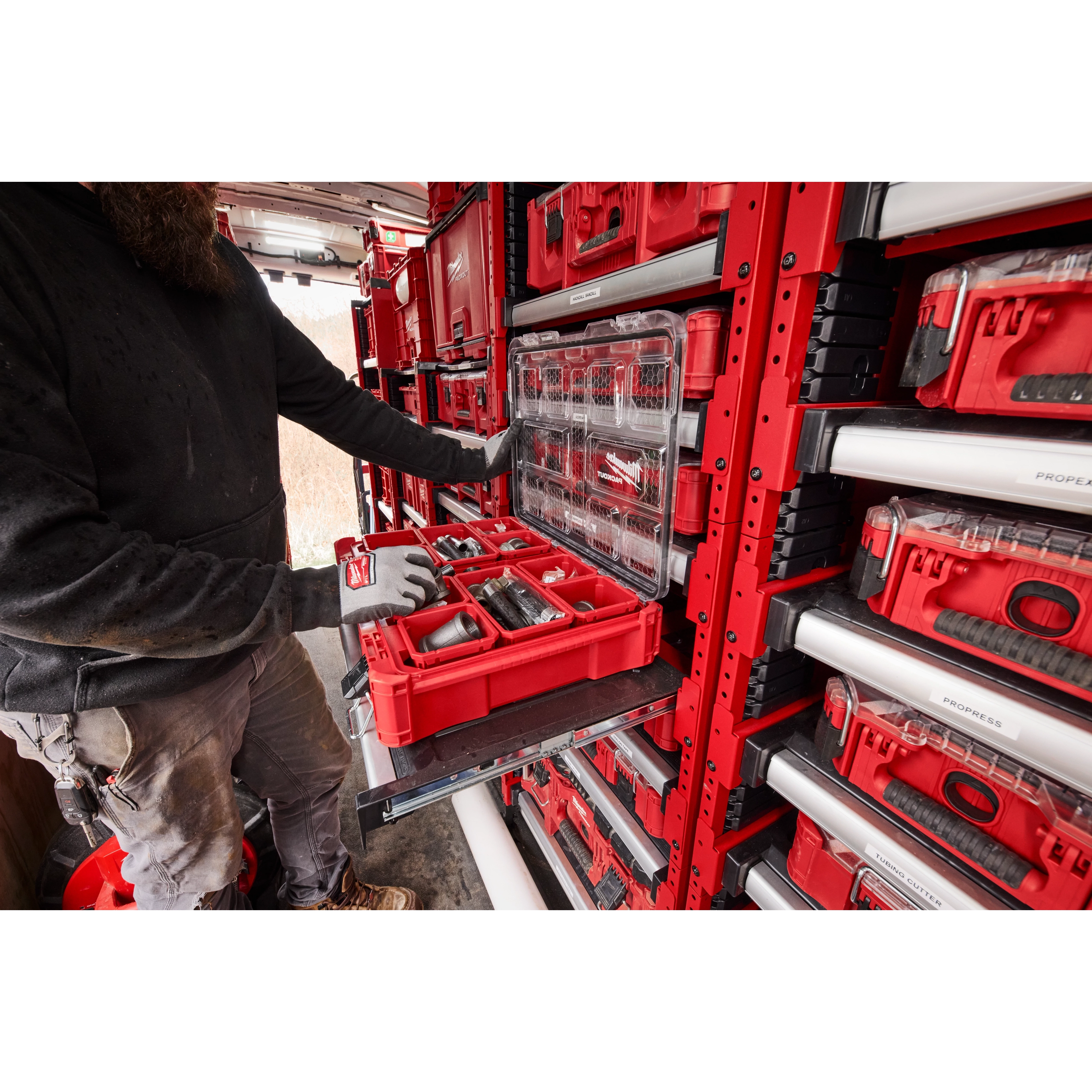 The PACKOUT™ Rack Kit is shown in use. A person in workwear retrieves a red, compartmentalized toolbox from the rack system installed in a work van. The rack organizes multiple red and gray toolboxes efficiently against the walls.